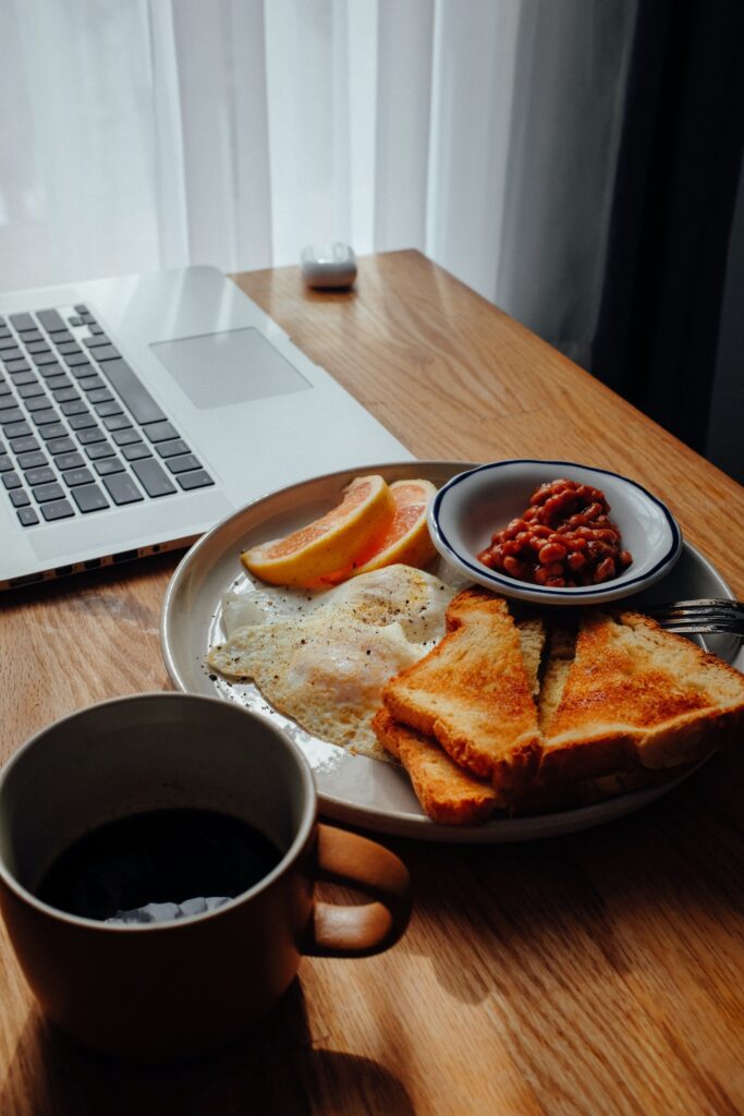 A plate of breakfast with toast, eggs, sliced oranges, and beans with a cup of coffee next to a laptop on a desk